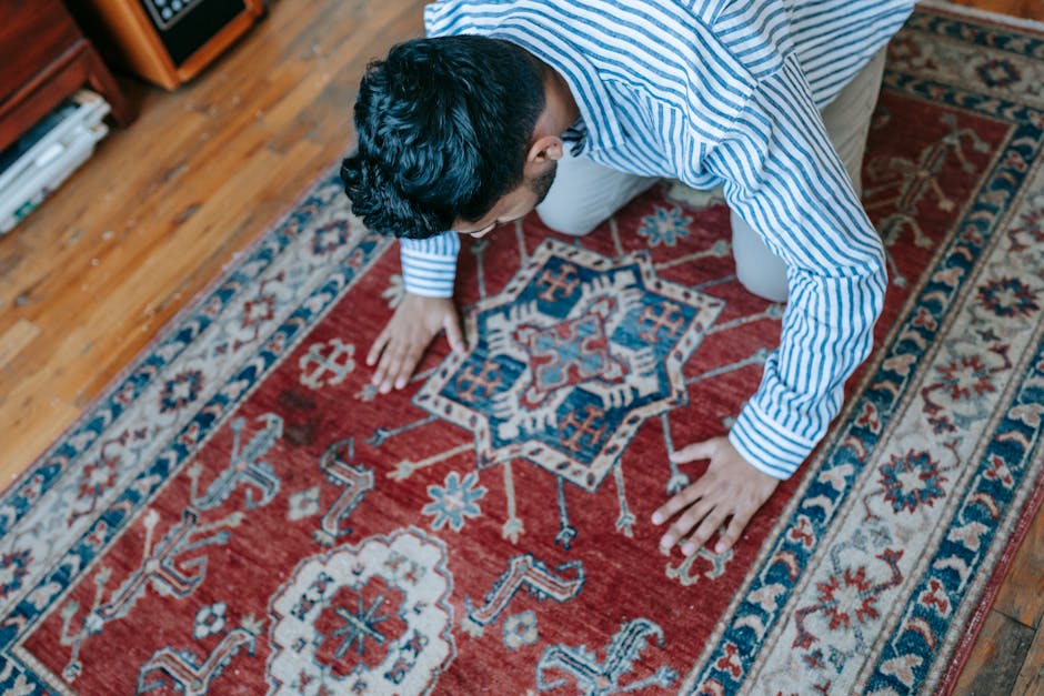A variety of colorful yarn balls in shades of pink, blue, green, yellow, purple, black, and gray are scattered on a patterned red and blue Persian-style rug, with a vintage silver pitcher placed among